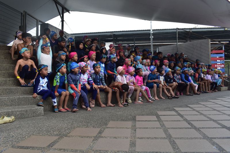 A big group of young swimmers in caps and goggles sitting together on the steps beside the pool