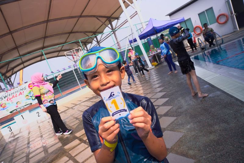 A smiling swimmer in goggles holding up a first-place Swimming Gala card