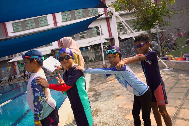 Four young swimmers in a line by the pool, hands on each other's shoulders, smiling at the camera